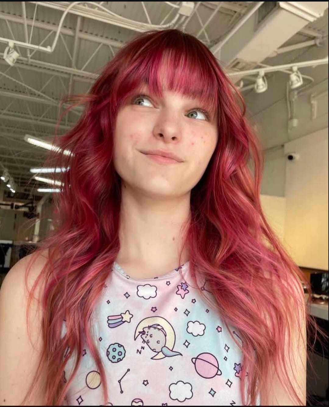 Young woman with vibrant red hair and a pastel unicorn-themed shirt in a modern room.