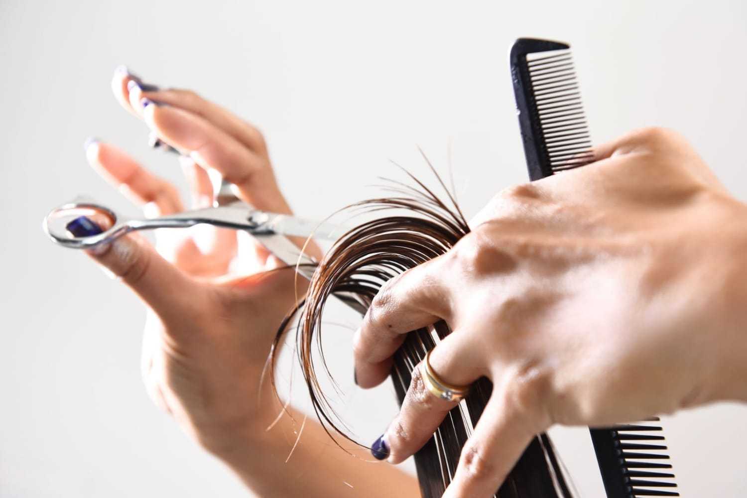 Hands trimming hair with scissors and comb against a plain background.